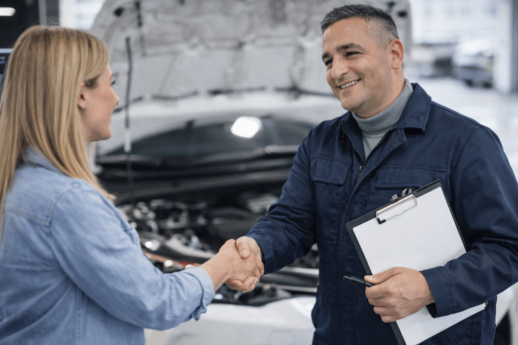 Ein Mechaniker mit Klemmbrett lächelt und schüttelt einer Frau nach Übergabe eines Kostenvoranschlags vor offener Motorhaube die Hand
